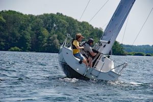 Das Bild zeigt zwei Personen auf einem Segelboot, das auf einem See gleitet. Das Boot ist leicht geneigt, was darauf hindeutet, dass der Wind die Segel füllt und für Vortrieb sorgt. Die beiden Personen tragen Freizeitkleidung und scheinen aktiv am Segeln beteiligt zu sein. Auf der Seite des Bootes steht der Name **„Hedda“**, was ihm eine persönliche Note verleiht. Im Hintergrund erstrecken sich Bäume und eine bewaldete Uferlinie, die eine friedliche Atmosphäre schaffen. Die Szene vermittelt Dynamik, Freiheit und die Freude am Wassersport inmitten einer schönen Naturkulisse.