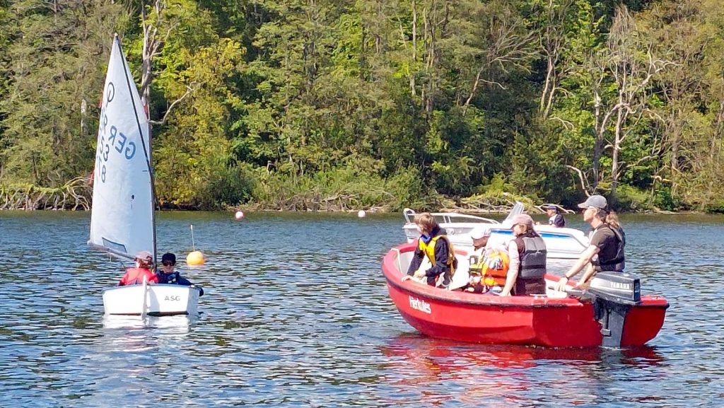 Kleines Segelboot und rotes Motorboot auf ruhigem See, umgeben von dichtem Wald. Menschen mit Schwimmwesten üben Manöver, orange Bojen markieren den Bereich, entspannte Trainingsatmosphäre auf dem Wasser.