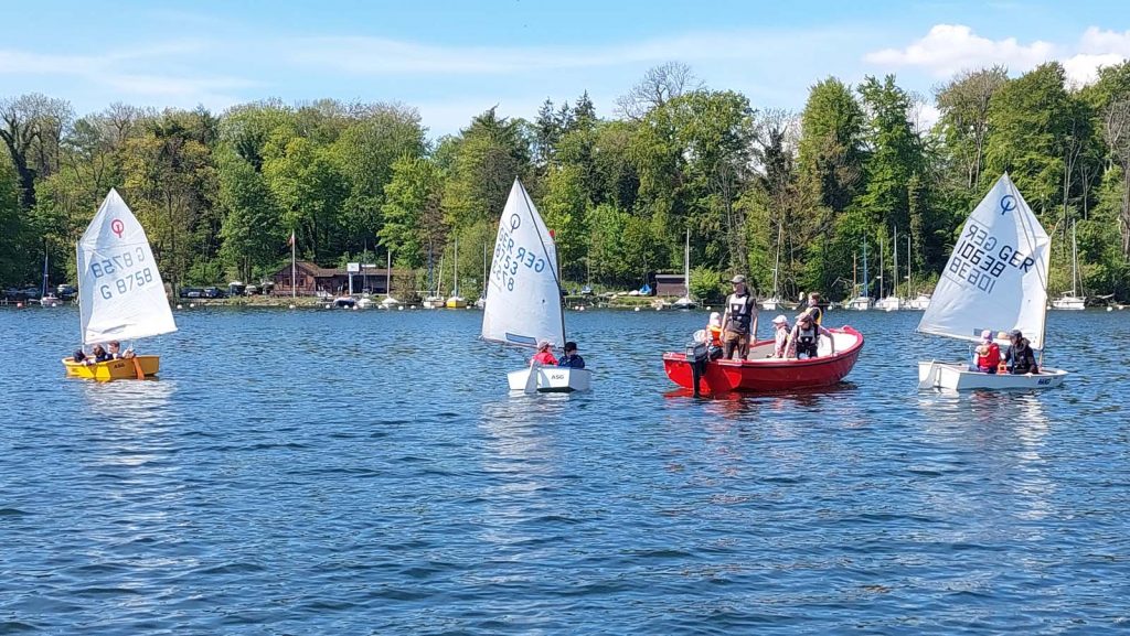 Mehrere kleine Segelboote auf ruhigem See, Segler mit Schwimmwesten, klarer Himmel, rotes Motornoot „Herkules“ im Vordergrund, weitere Jollen im Hintergrund, sommerliche Trainings‑ und Freizeitstimmung auf dem Wasser.