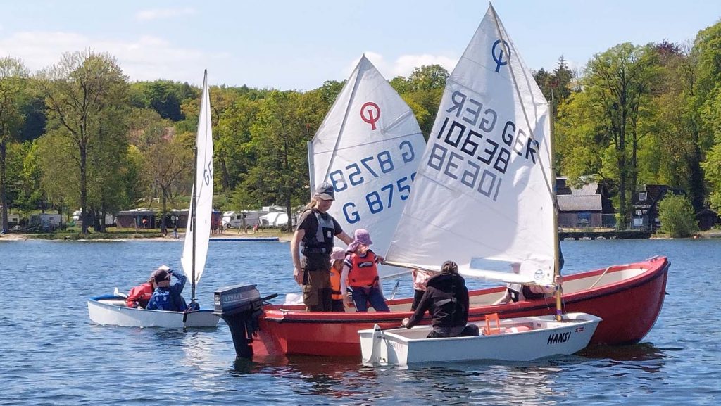 Kleines Segelboot und rotes Motorboot auf ruhigem See, umgeben von dichtem Wald. Menschen mit Schwimmwesten üben Manöver, orange Bojen markieren den Bereich, entspannte Trainingsatmosphäre auf dem Wasser.
