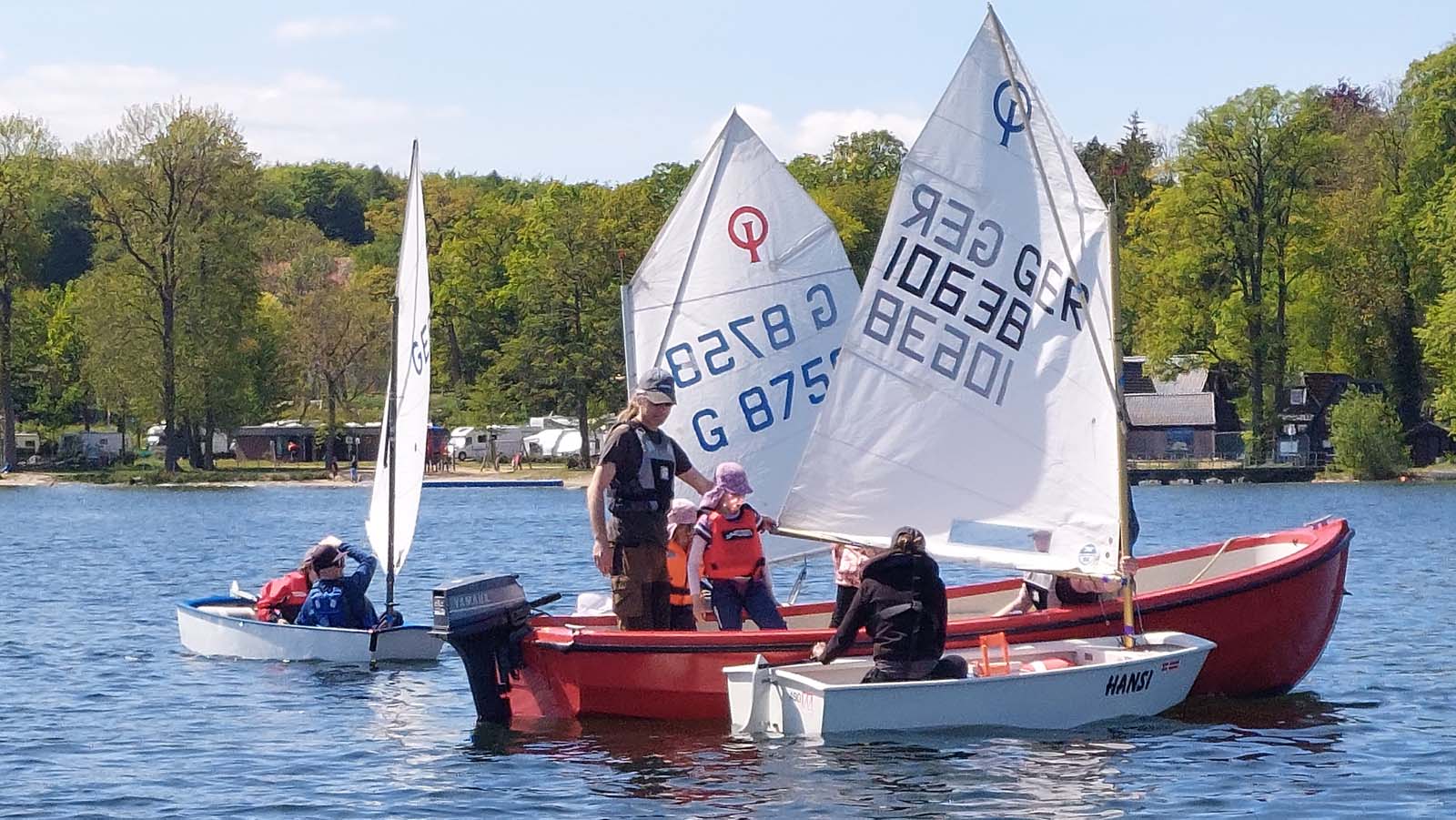 Kleines Segelboot und rotes Motorboot auf ruhigem See, umgeben von dichtem Wald. Menschen mit Schwimmwesten üben Manöver, orange Bojen markieren den Bereich, entspannte Trainingsatmosphäre auf dem Wasser.