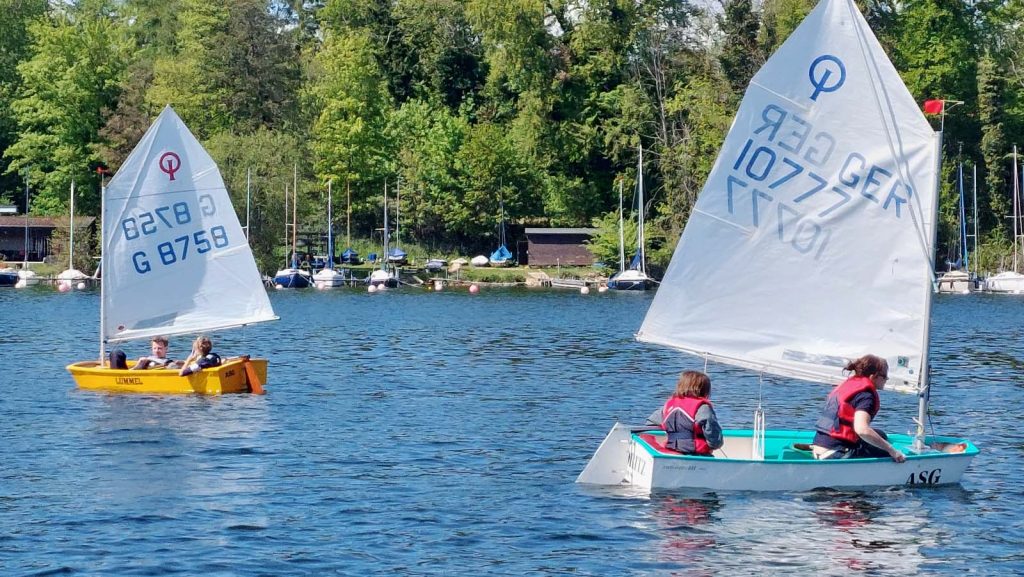 Ruhige Segelszene auf kleinem Gewässer: Zwei Jollen mit Seglern im Vordergrund, weitere Boote am Steg, grünes Ufer, Bäume, Bootshaus und entspannte Atmosphäre eines Trainings oder Vereinsnachmittags.