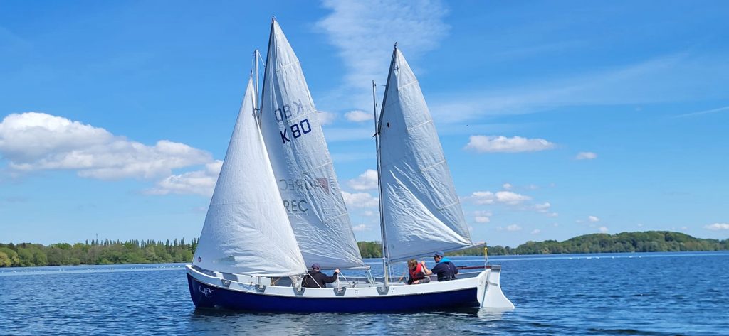 Segelkutter mit dreiweißen Segeln gleitet über ruhiges Wasser, drei Personen an Bord, dahinter weiteres Boot und bewaldetes Ufer, friedliche Stimmung unter leicht bewölktem Himmel.