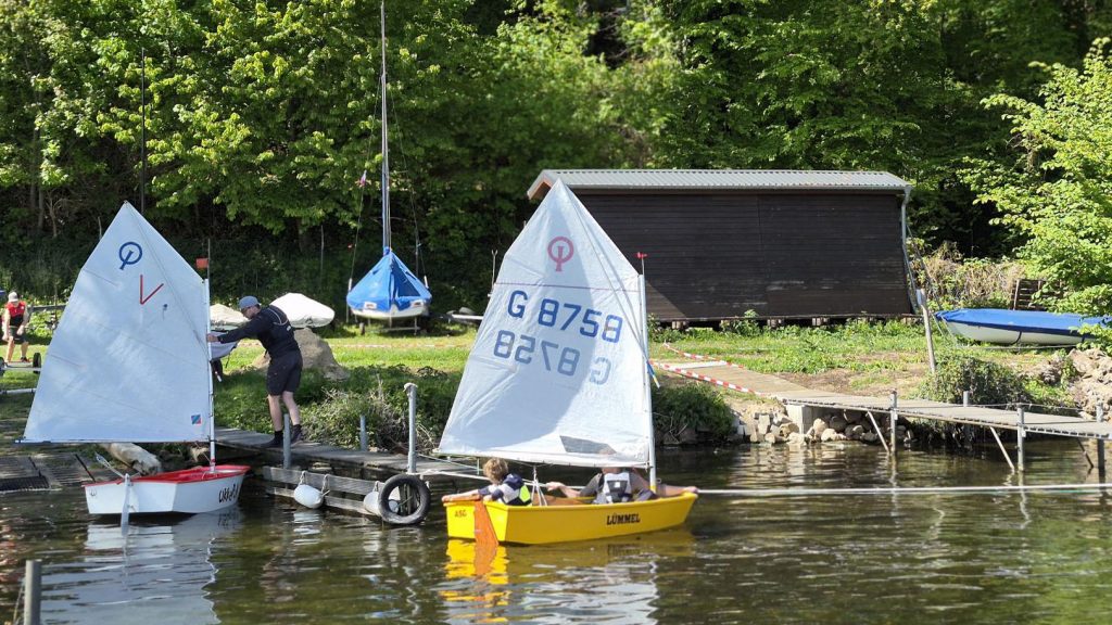 Kleiner Segelhafen mit zwei Optimistenjollen im Vordergrund, Menschen bereiten Boote vor, ruhiges Wasser, grünes Ufer mit Bäumen, Bootsschuppen und gelagerten Booten, entspannte Trainings‑ oder Vereinsatmosphäre.