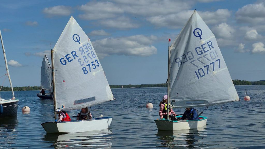 Zwei Optimistenjollen gleiten über ruhiges Wasser, junge Segler tragen Schwimmwesten, im Hintergrund weitere Boote und Bojen, darüber ein teils bewölkter Himmel, friedliche Trainingsszene auf dem See.