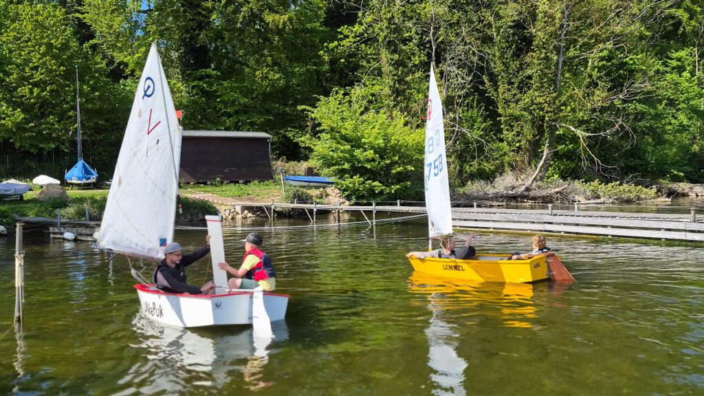 Kleiner Segelhafen mit zwei Optimistenjollen im Vordergrund, Menschen bereiten Boote vor, ruhiges Wasser, grünes Ufer mit Bäumen, Bootsschuppen und gelagerten Booten, entspannte Trainings‑ oder Vereinsatmosphäre.